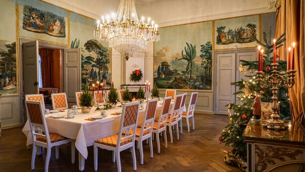 Une salle à manger avec une longue table dressée pour un repas, entourée de chaises, un lustre au-dessus et un sapin de Noël sur le côté.