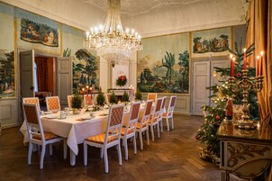 Ein Esszimmer mit langem Tisch, gedeckt für eine Mahlzeit, umgeben von Stühlen, mit einer darüber hängenden Kronleuchter und einer Weihnachtstanne seitlich.