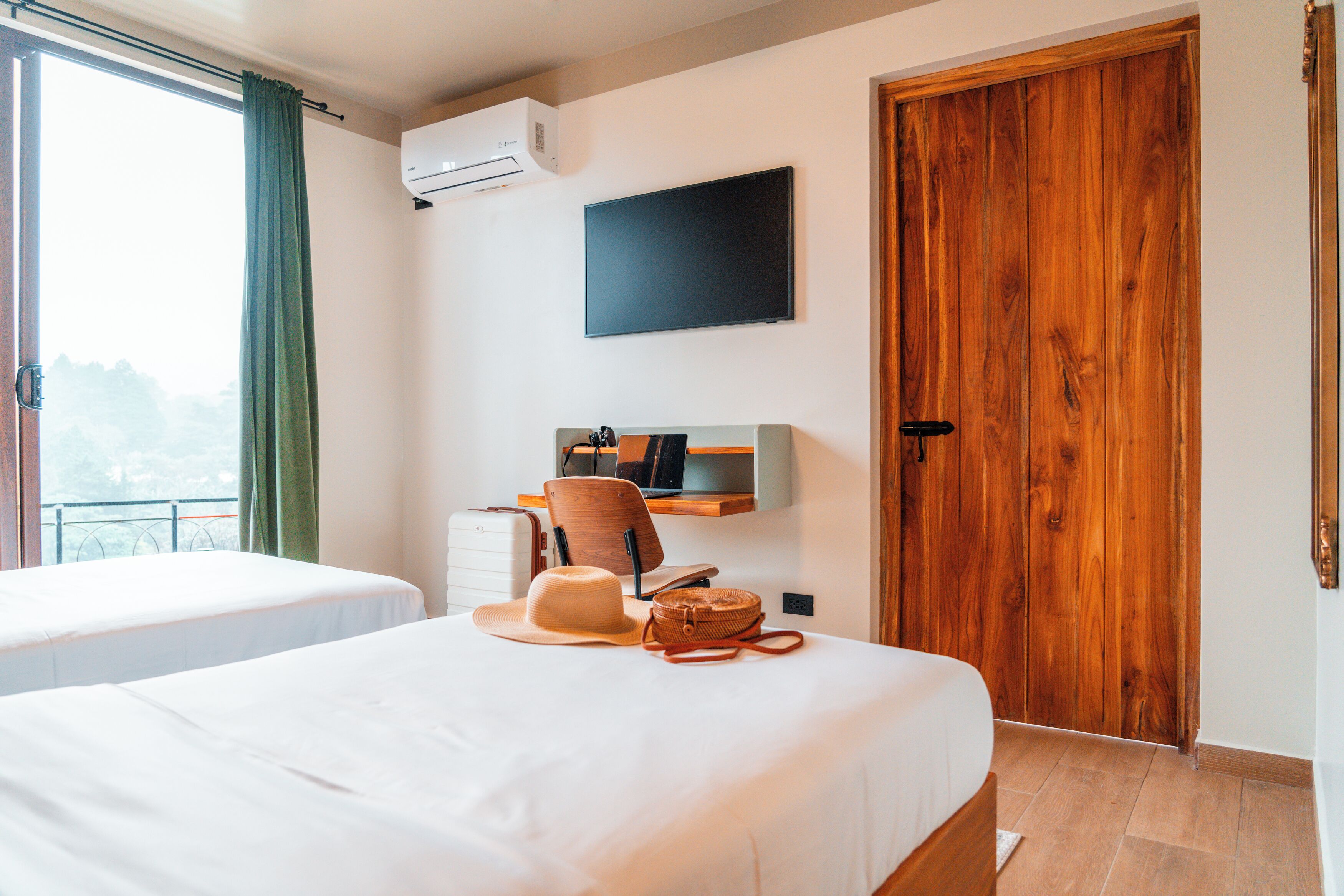 A hotel room with two beds, a TV, a wooden door, and a window with curtains.
