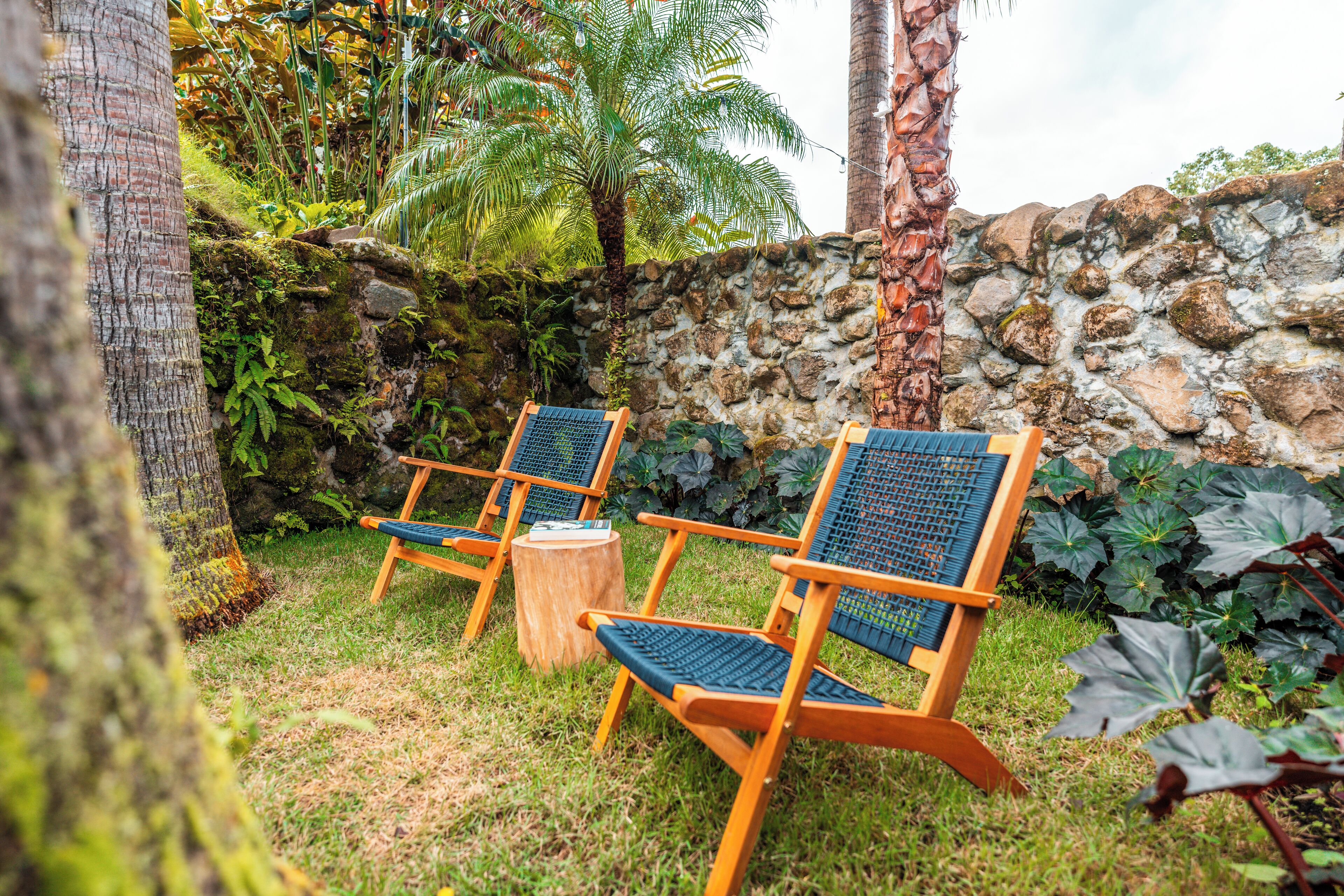Two wooden chairs with blue mesh backrests and seats, placed on grass with a stone wall and tropical plants in the background.