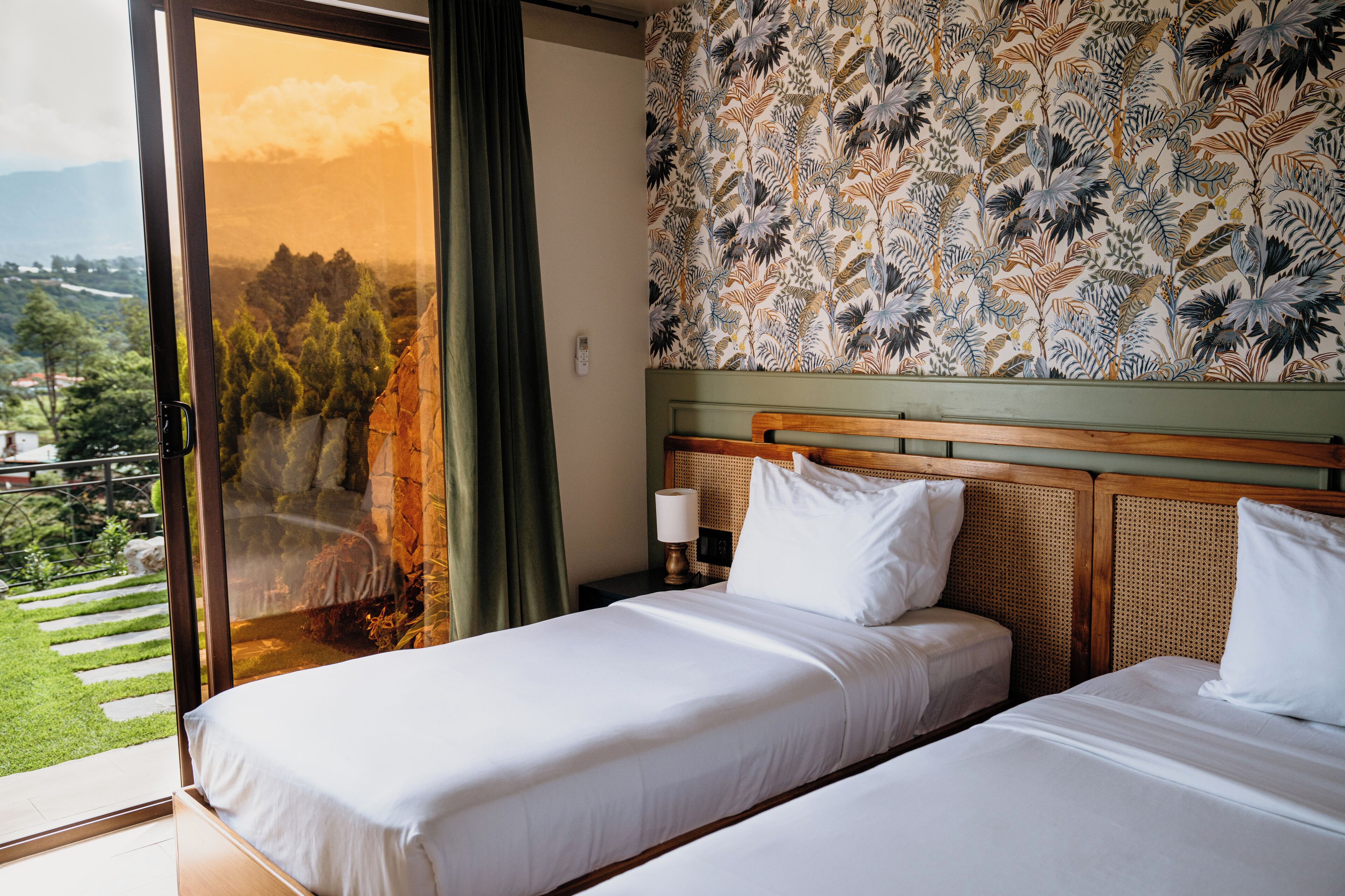 Habitación de hotel con dos camas, un ventanal con vistas a un jardín y montañas, y una pared con diseño.