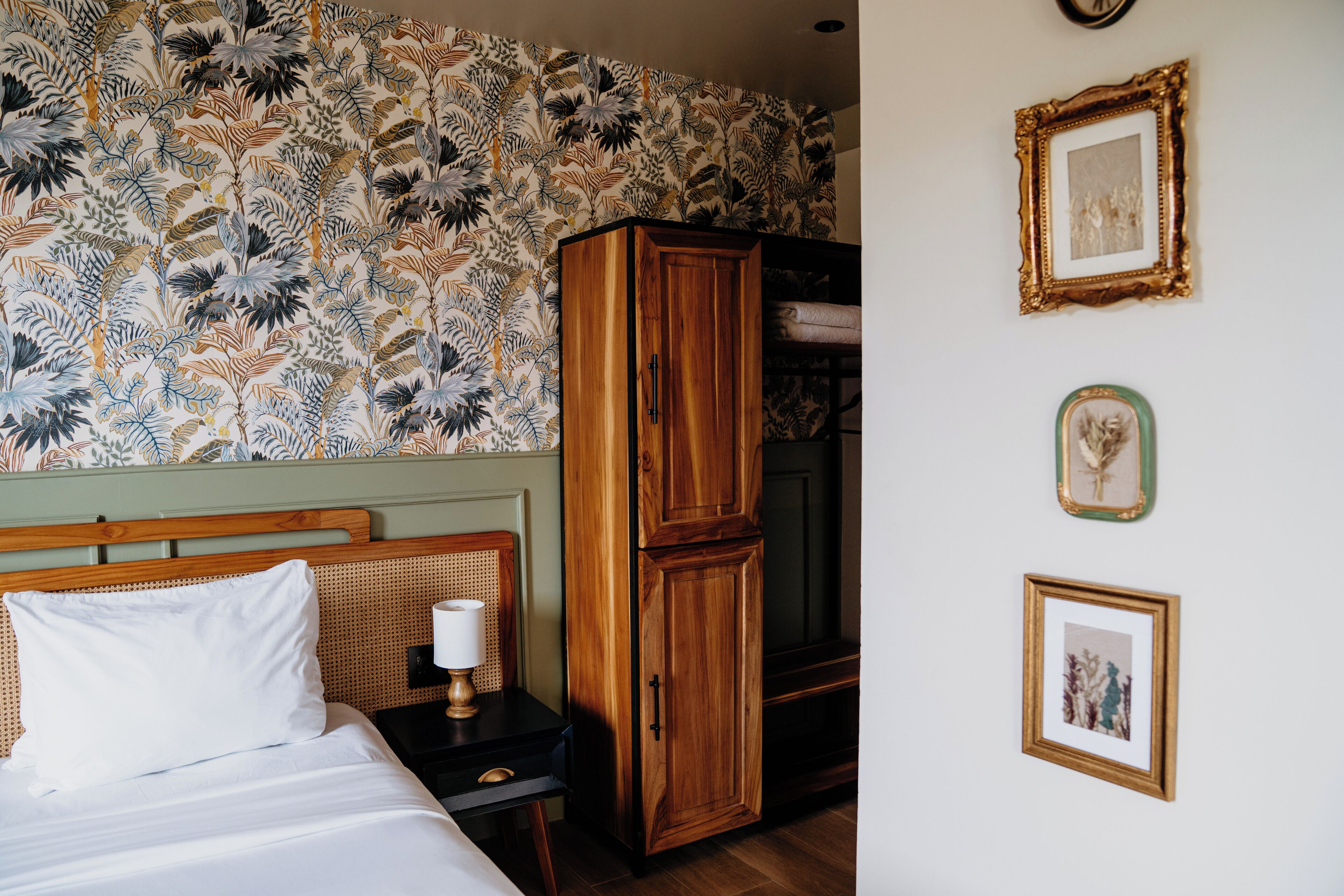 Een hotelkamer met een bed, een houten garderobe, een nachtkastje met een lamp, en een wand met bloemenbehang en ingelijste foto's.