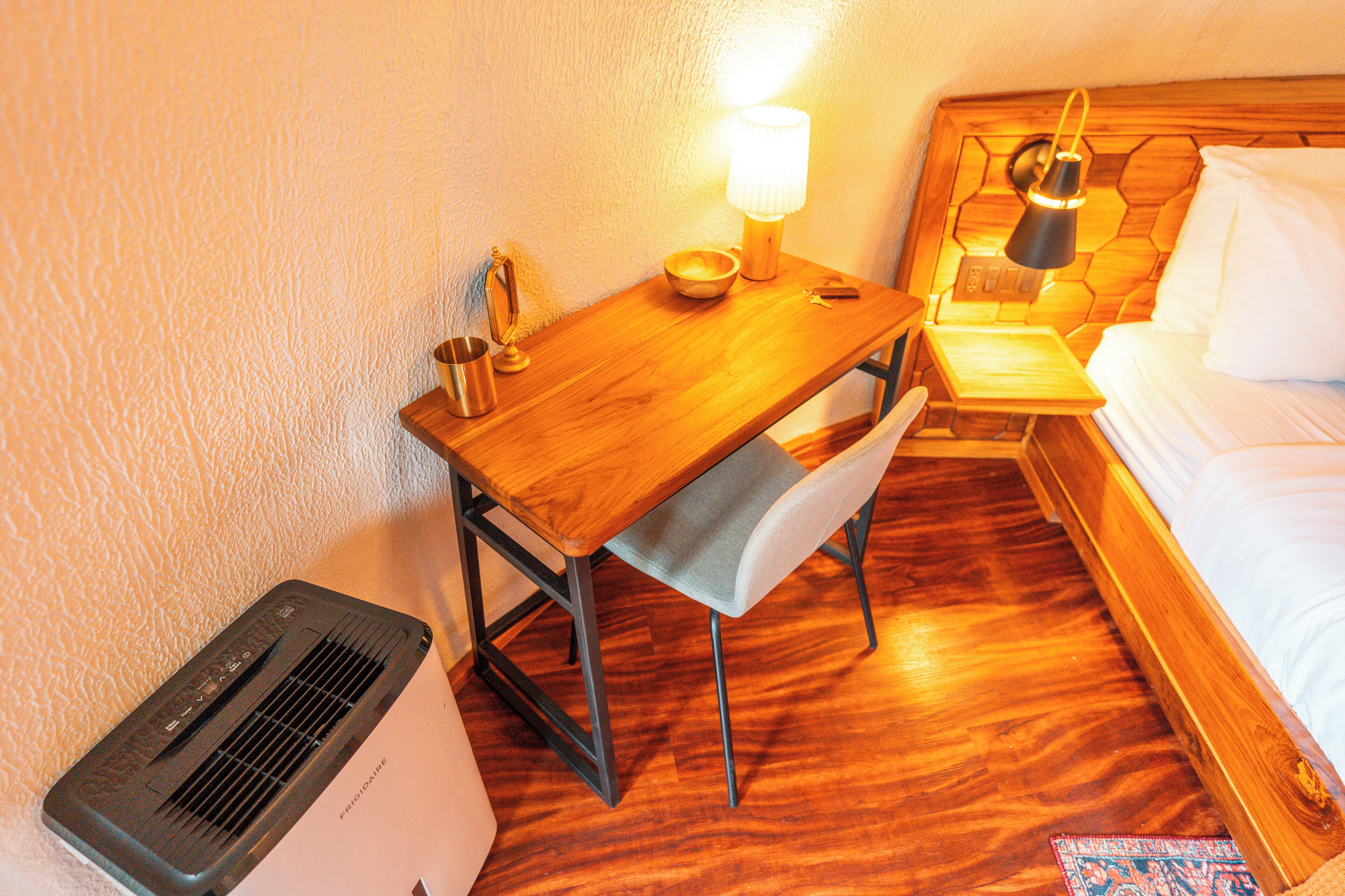A hotel room with a wooden desk, a chair, a bedside table with a lamp, a bed, and a heater.