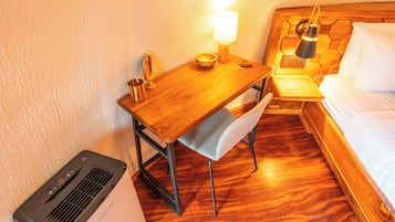 A hotel room with a wooden desk, a chair, a bedside table with a lamp, a bed, and a heater.