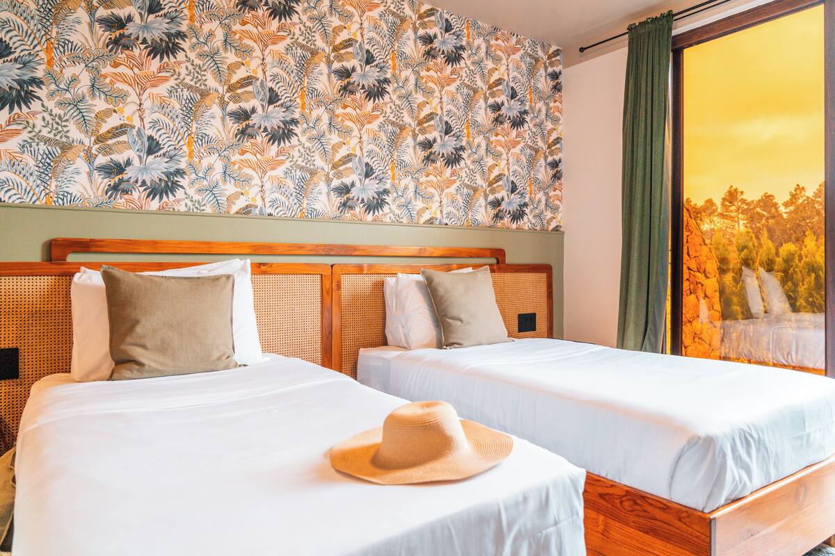 A hotel room with two beds, a floral wallpaper, and a view of trees through a window.
