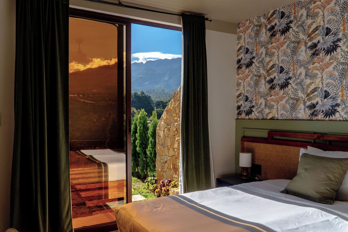 A hotel room with a bed, a nightstand, a lamp, and a view of mountains through a sliding door.