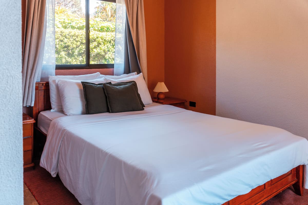 A neatly made bed with white and green pillows, a wooden nightstand with a lamp, and a window with curtains.