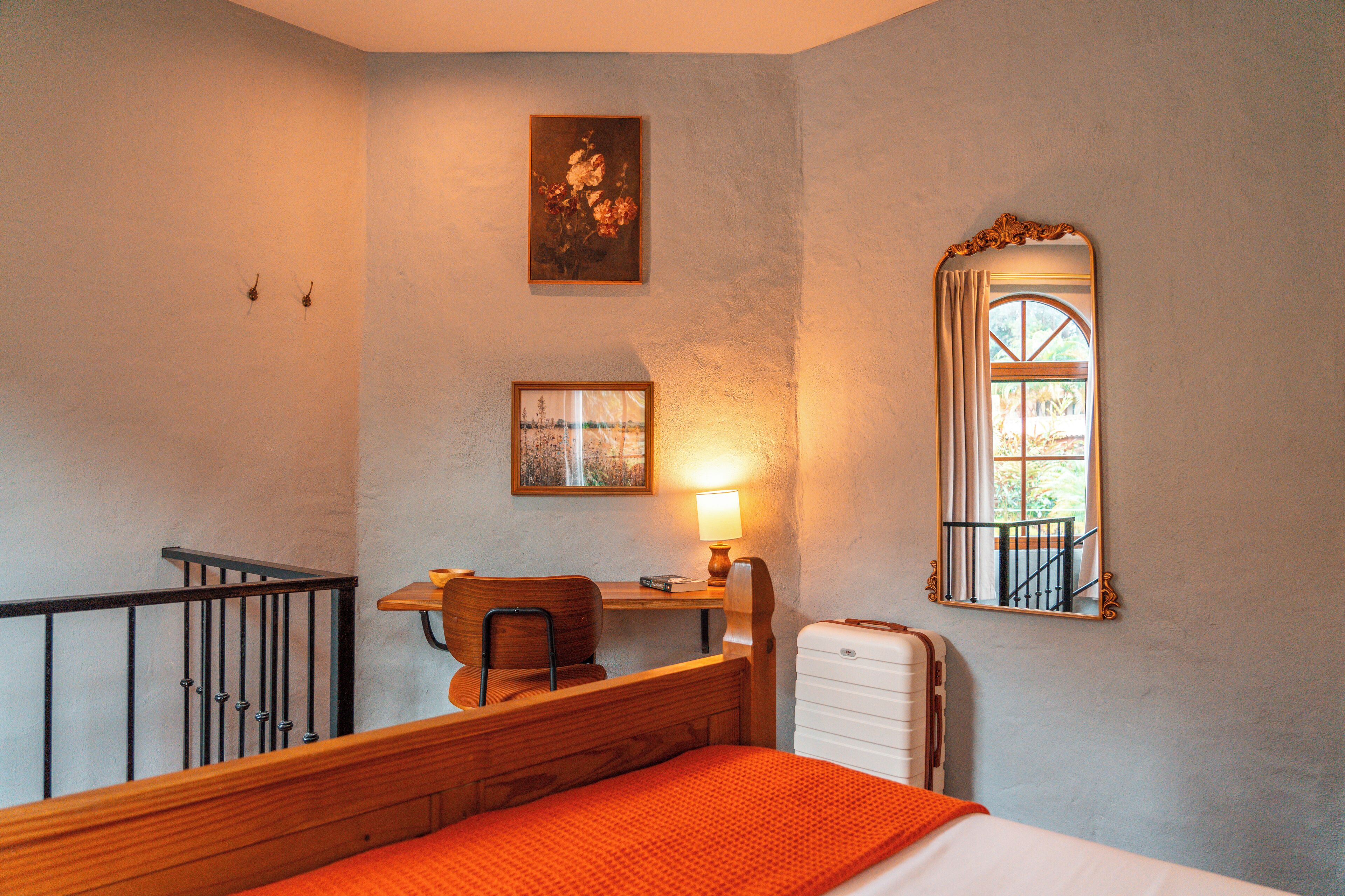 A bedroom with a bed, a desk with a chair, a lamp, a suitcase, and a window with a view of greenery.