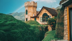 A castle-like building with a tower, surrounded by lush green hills and mountains in the background. - Bambuda Castle - Hostel (Los Naranjos)