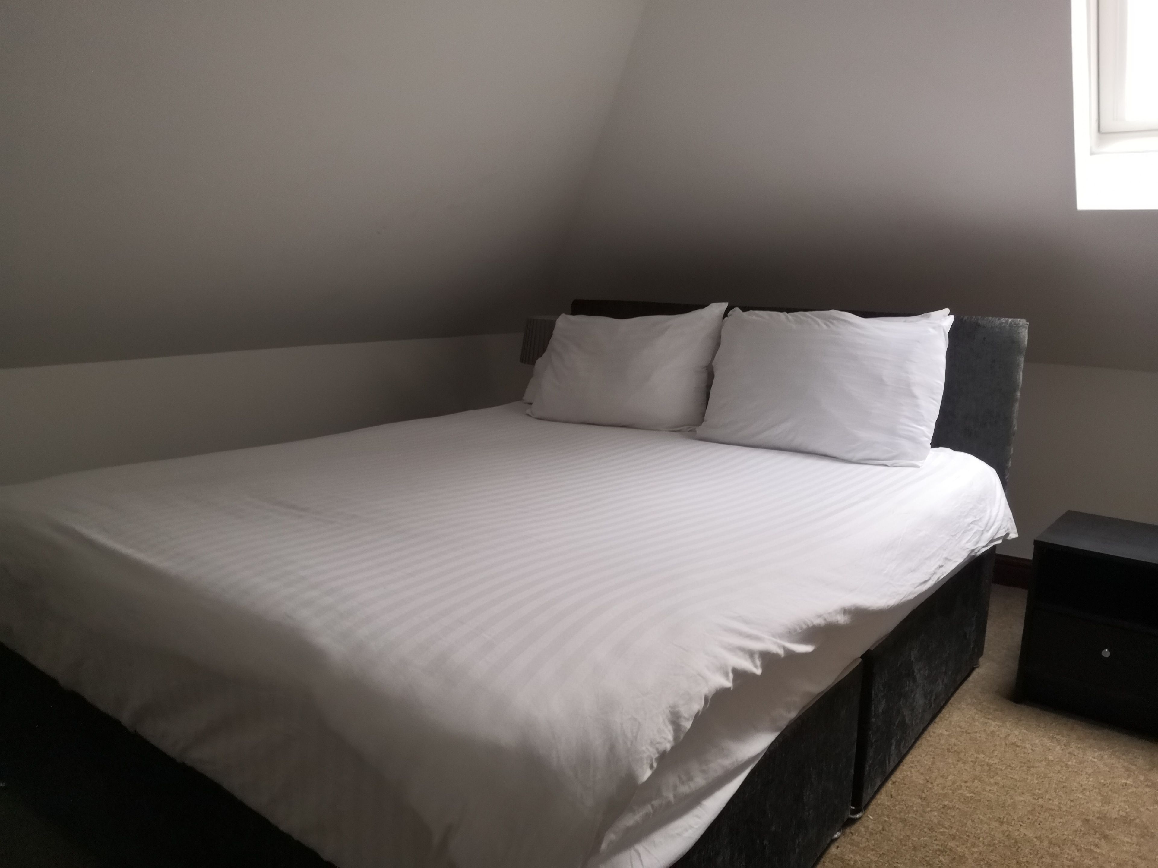 A bedroom with a bed, two pillows, a nightstand, and a window.