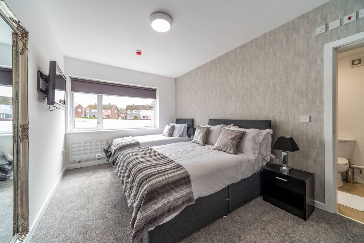 A modern bedroom with a large bed, a bedside table, a mirror, and a window with a view of houses.