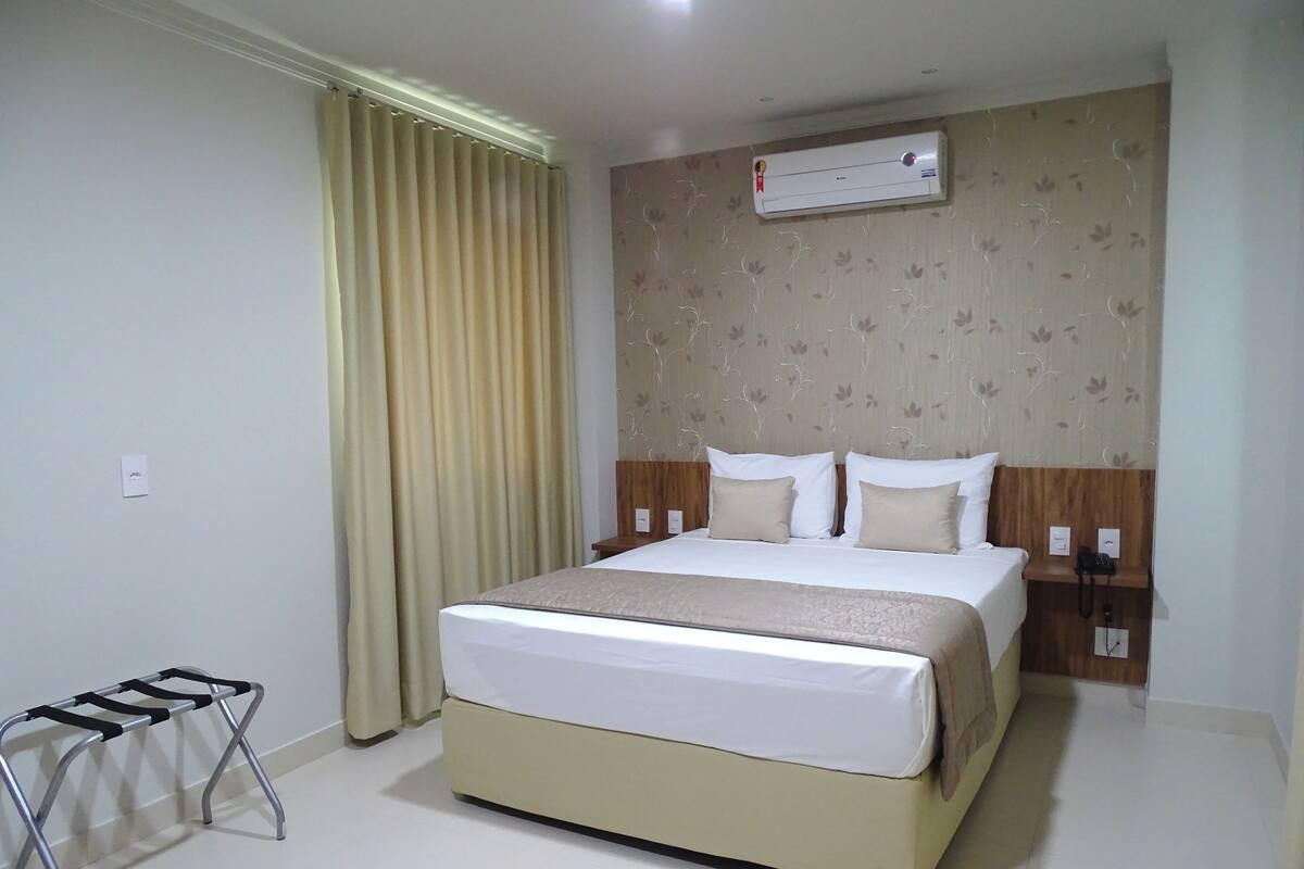 A hotel room with a bed, curtains, an air conditioning unit, a bedside table with a phone, and a small table with a towel rack.