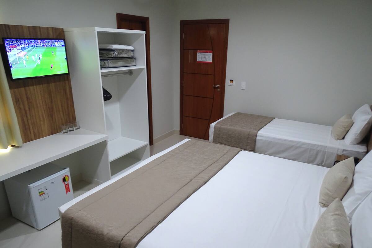 A hotel room with two beds, a TV mounted on the wall, a wardrobe, and a small refrigerator.