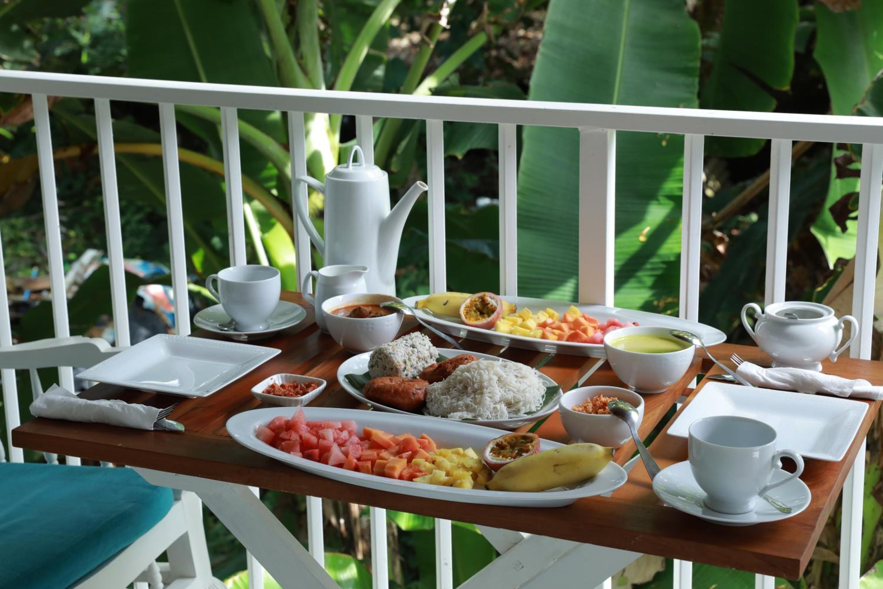 A wooden table set with various dishes, including rice, vegetables, and a banana, on a balcony with a white railing and lush greenery in the background.