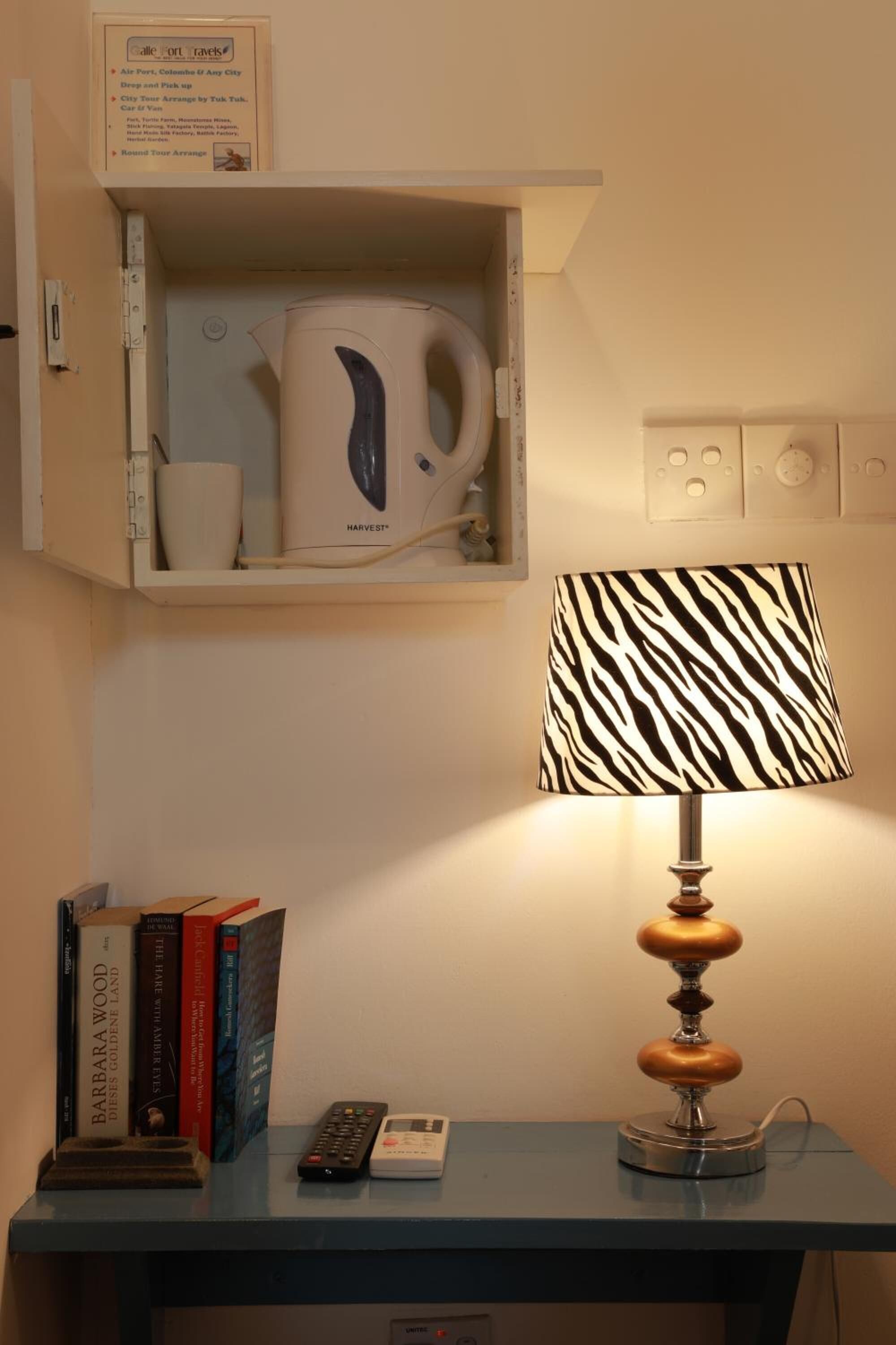 Ein Zimmer mit einer Tischlampe, einem Regal mit Büchern und einer Fernbedienung sowie einem Schrank mit Wasserkocher und Tassen.