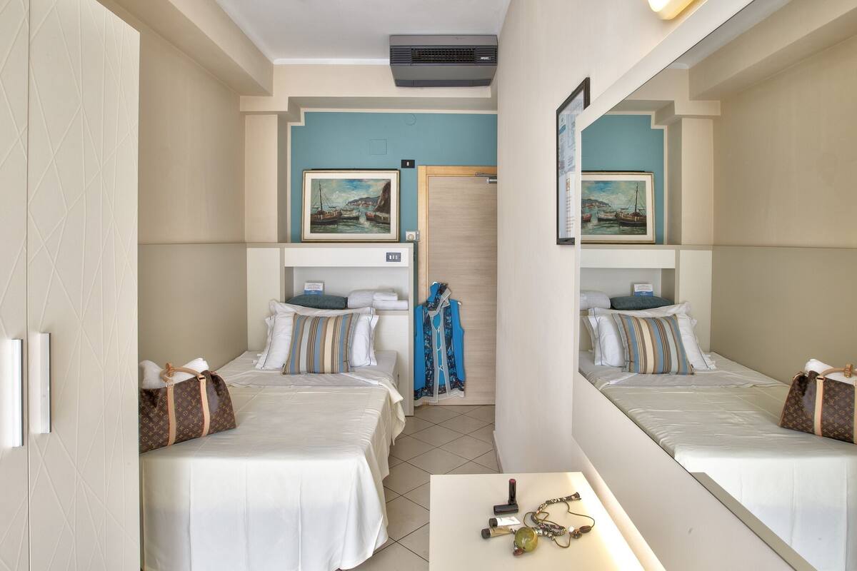 Habitación de hotel con dos camas, una mesita y un espejo. Hay cuadros en las paredes y una puerta con pomo.