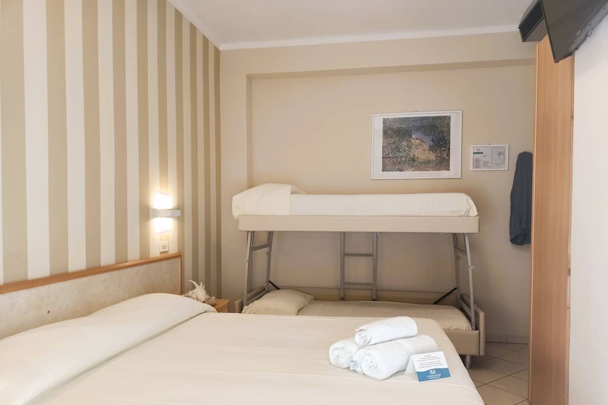 Habitación de hotel con literas, una cama individual y una mesita con lámpara. Se observa un cuadro en la pared y una toalla sobre la cama.