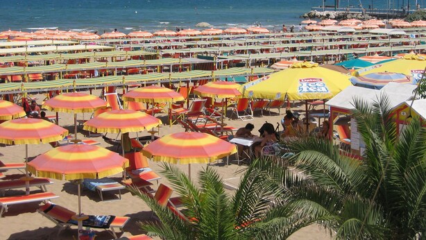 Una playa con numerosos sombrillas y sillas de playa, cielo despejado y el océano al fondo.