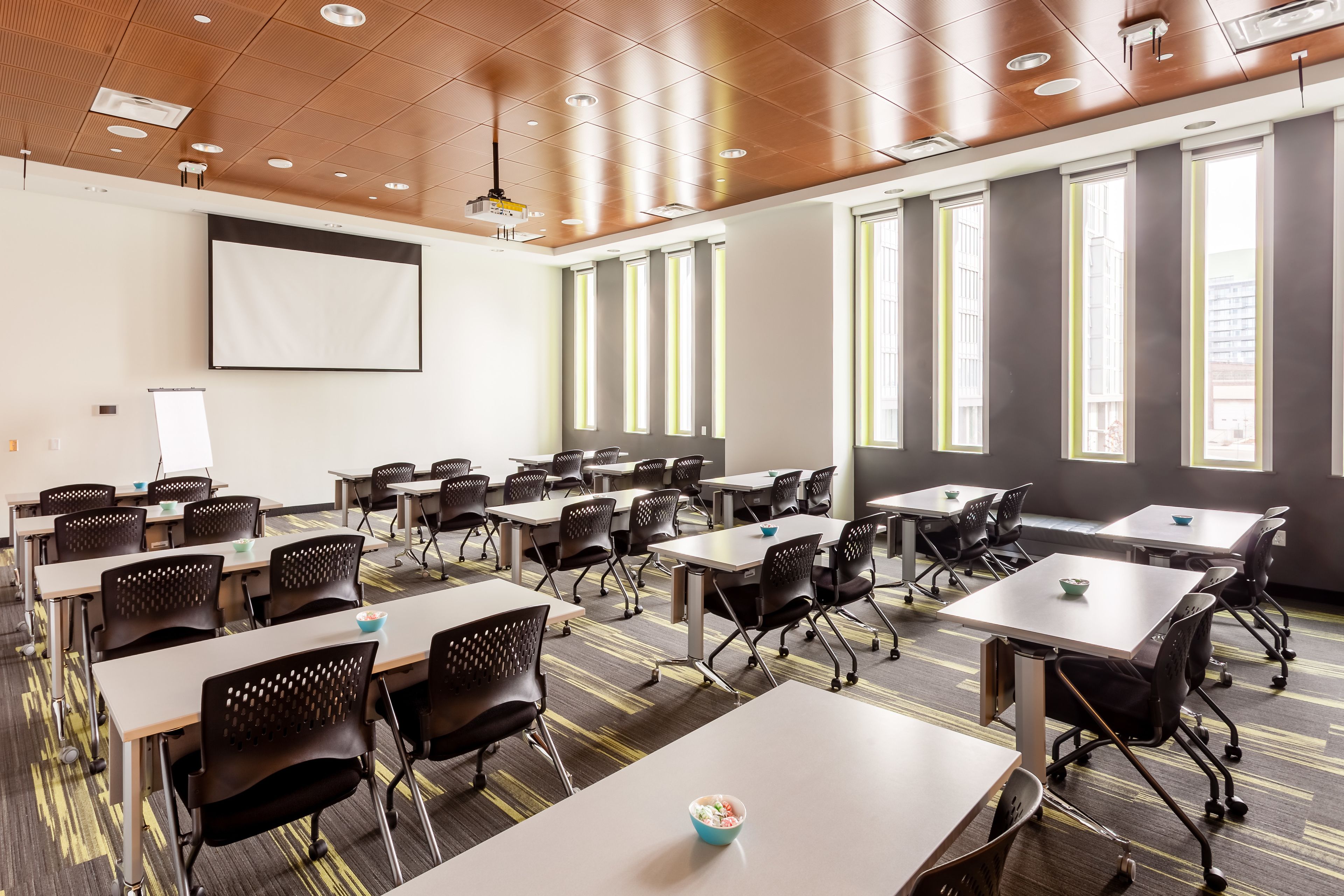 Un salón de conferencias con varias mesas y sillas, ventanas amplias y una pantalla de proyector.