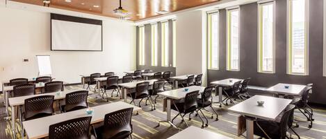 Un salón de conferencias con varias mesas y sillas, ventanas amplias y una pantalla de proyector.