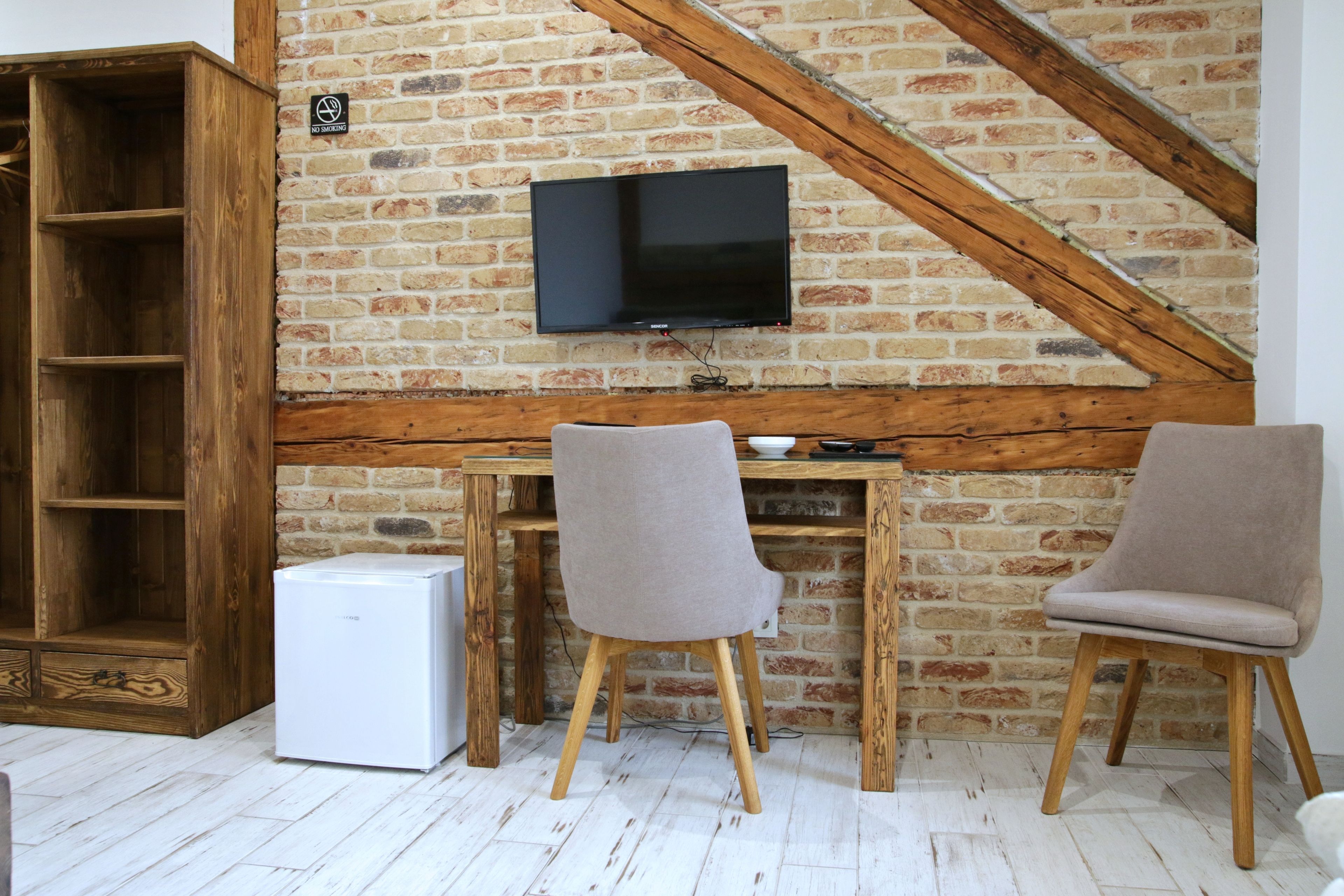 A room with a brick wall, a TV mounted on it, a wooden desk, two chairs, and a white refrigerator.