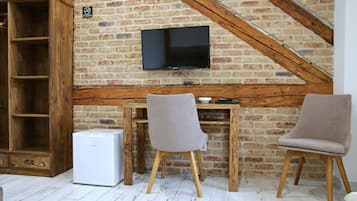 A room with a brick wall, a TV mounted on it, a wooden desk, two chairs, and a white refrigerator.