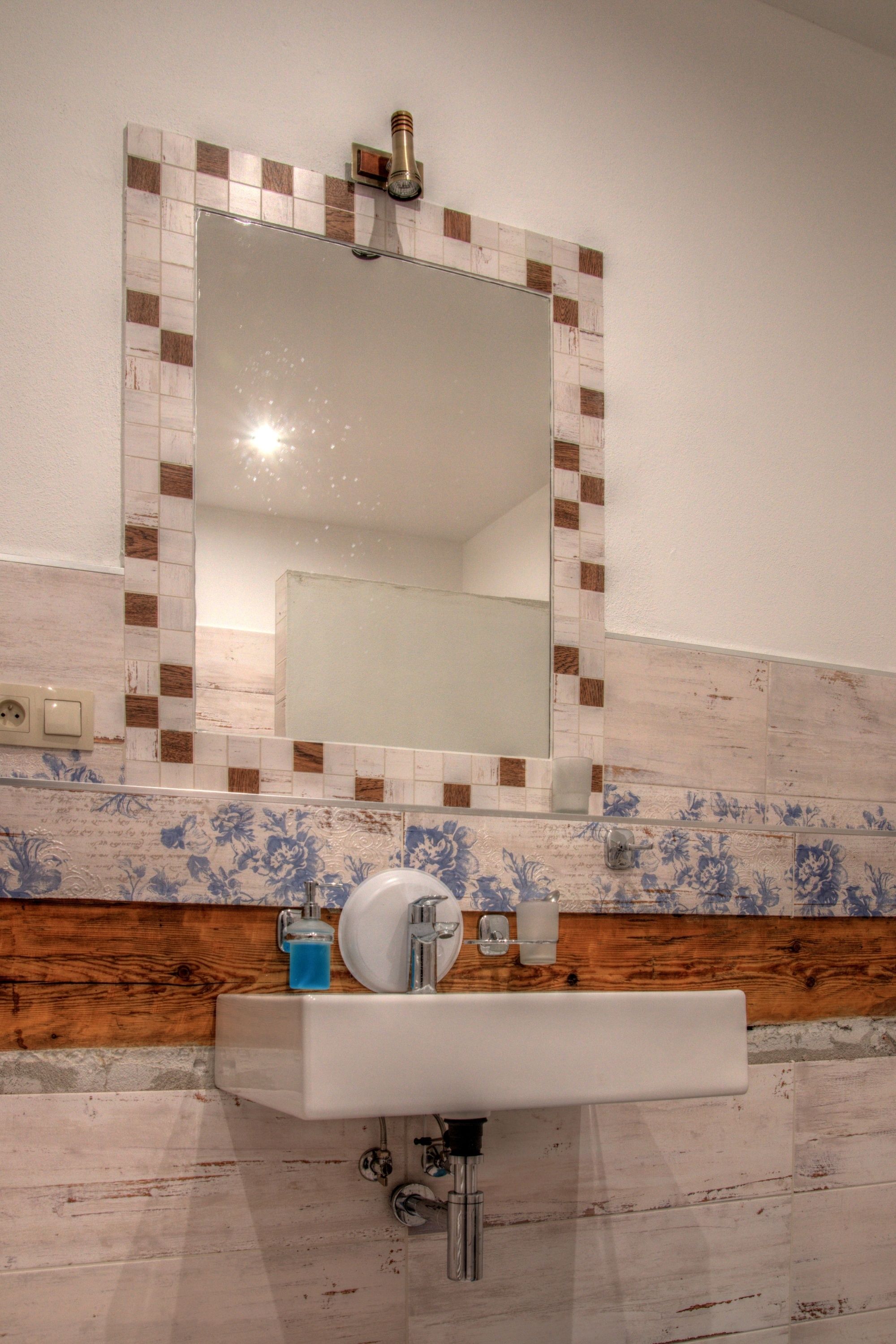 Un baño con lavamanos blanco, un espejo con borde de azulejos marrones y blancos, y azulejos florales azules en la pared.