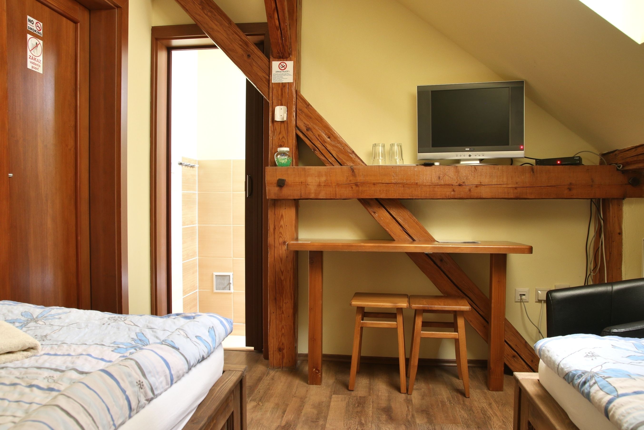 Une pièce avec un lit en bois, une petite table en bois, un téléviseur posé sur une étagère en bois, et une salle de bain visible à travers une porte ouverte.