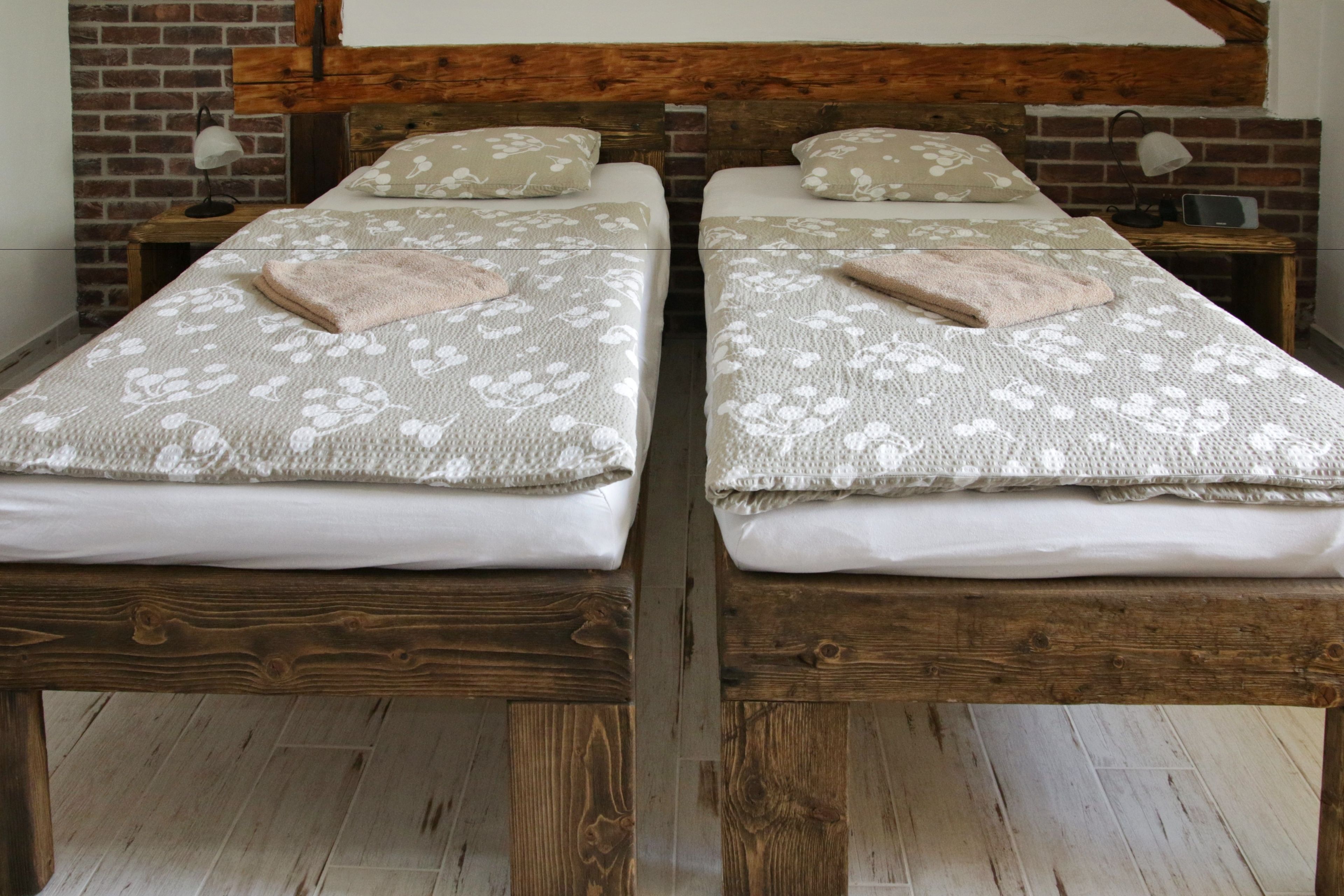 A room with two wooden beds, each with a floral-patterned bedspread and a folded towel. There is a brick wall and a wooden beam ceiling.
