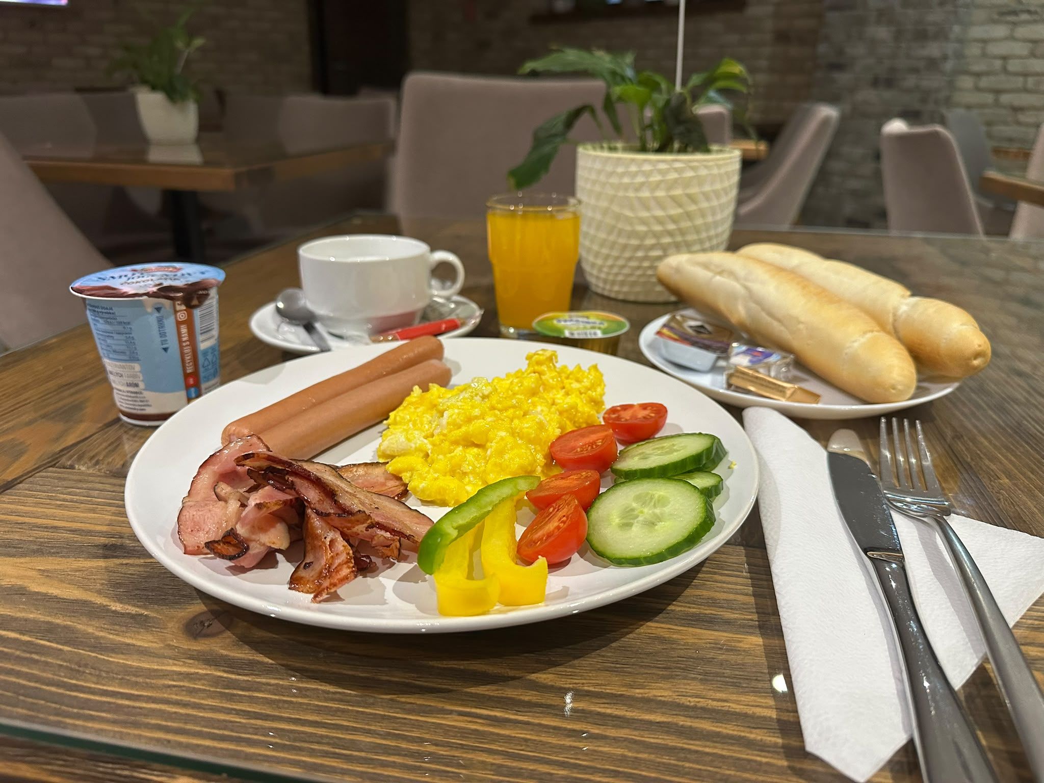 A breakfast plate with eggs, bacon, tomatoes, cucumbers, and bread rolls on a wooden table.