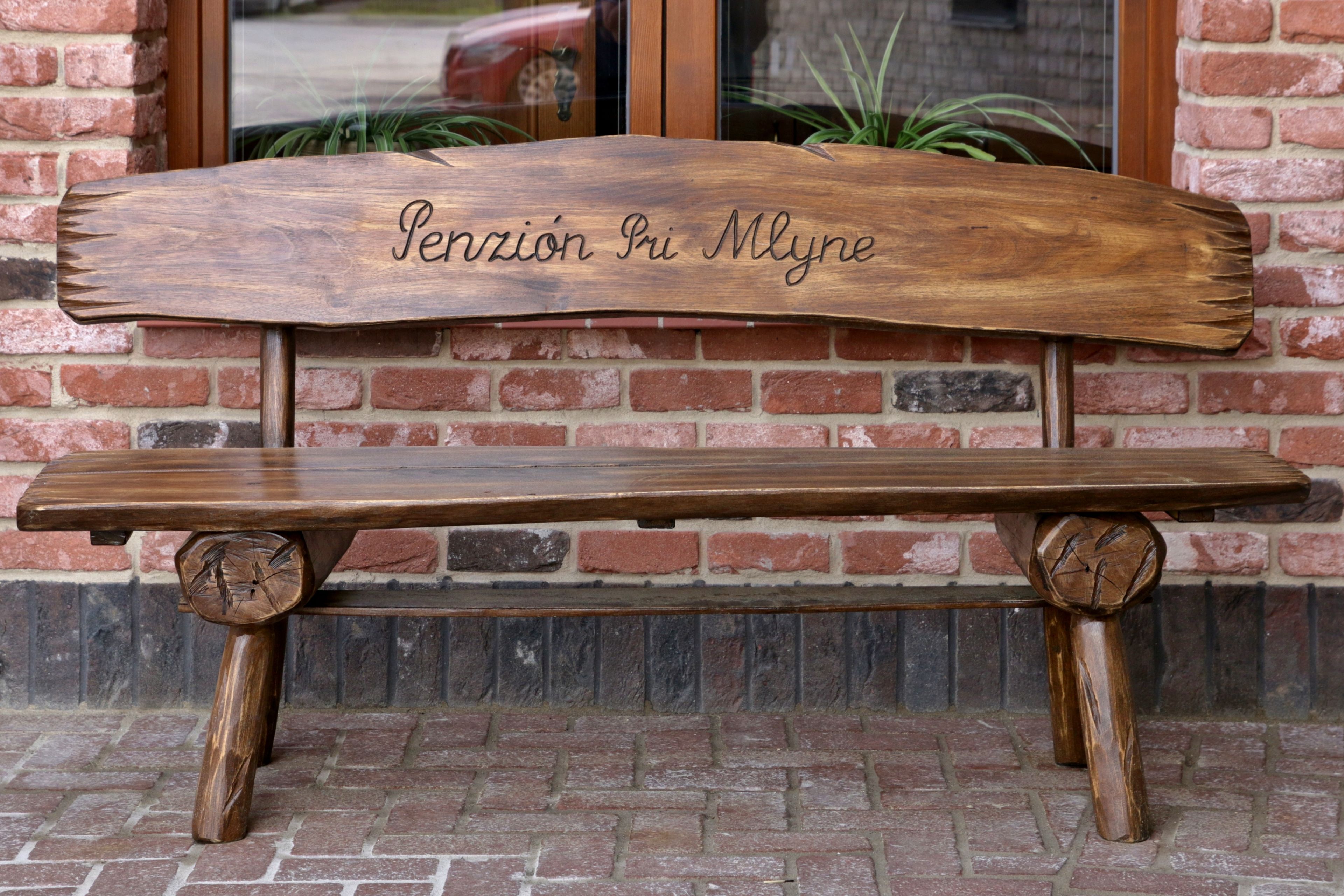A wooden bench with a carved backrest and the inscription "Pension Piu Mlyne" against a brick wall.