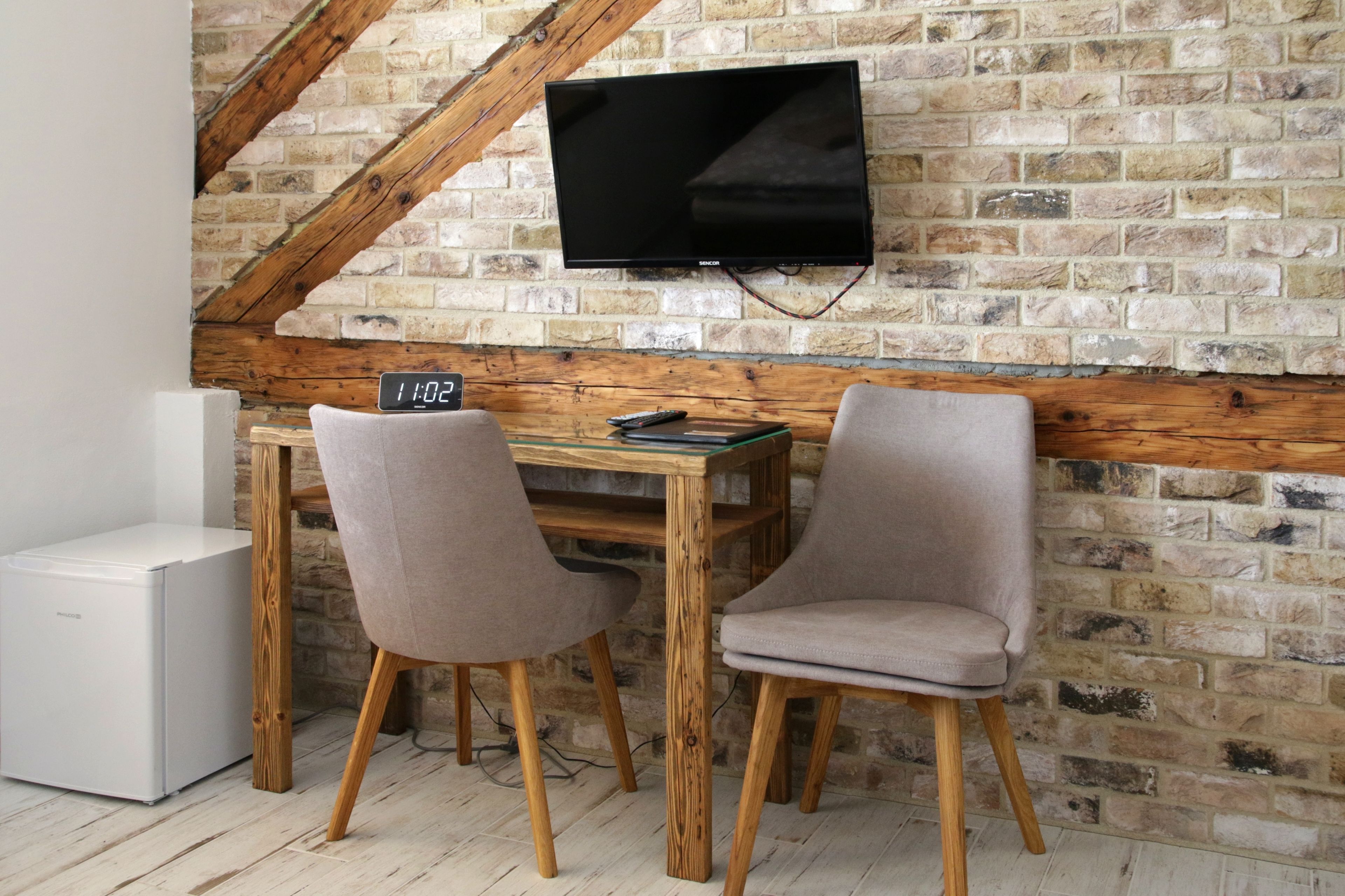Una habitación con pared de ladrillo, vigas de madera, un televisor sobre una mesa de madera con dos sillas, y un electrodoméstico blanco en el suelo.