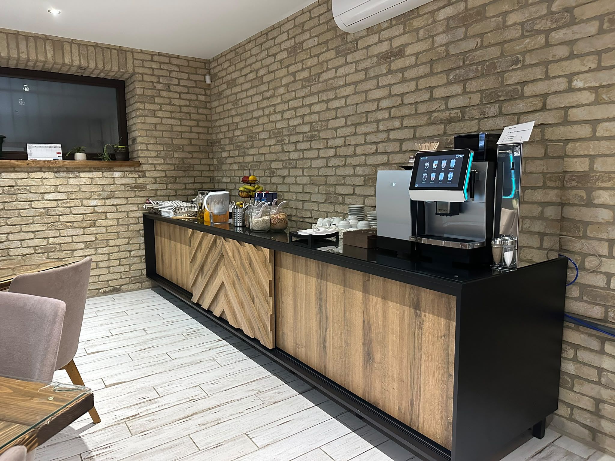 Zona de recepción de un hotel con pared de ladrillo, mostrador de recepción, máquina de café y área de descanso con sillas.