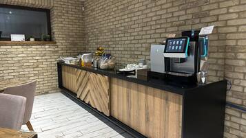 Zona de recepción de un hotel con pared de ladrillo, mostrador de recepción, máquina de café y área de descanso con sillas.