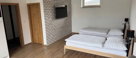 Une chambre avec un mur en briques, des poutres en bois, un lit avec une literie blanche, un canapé gris et un téléviseur fixé au mur.