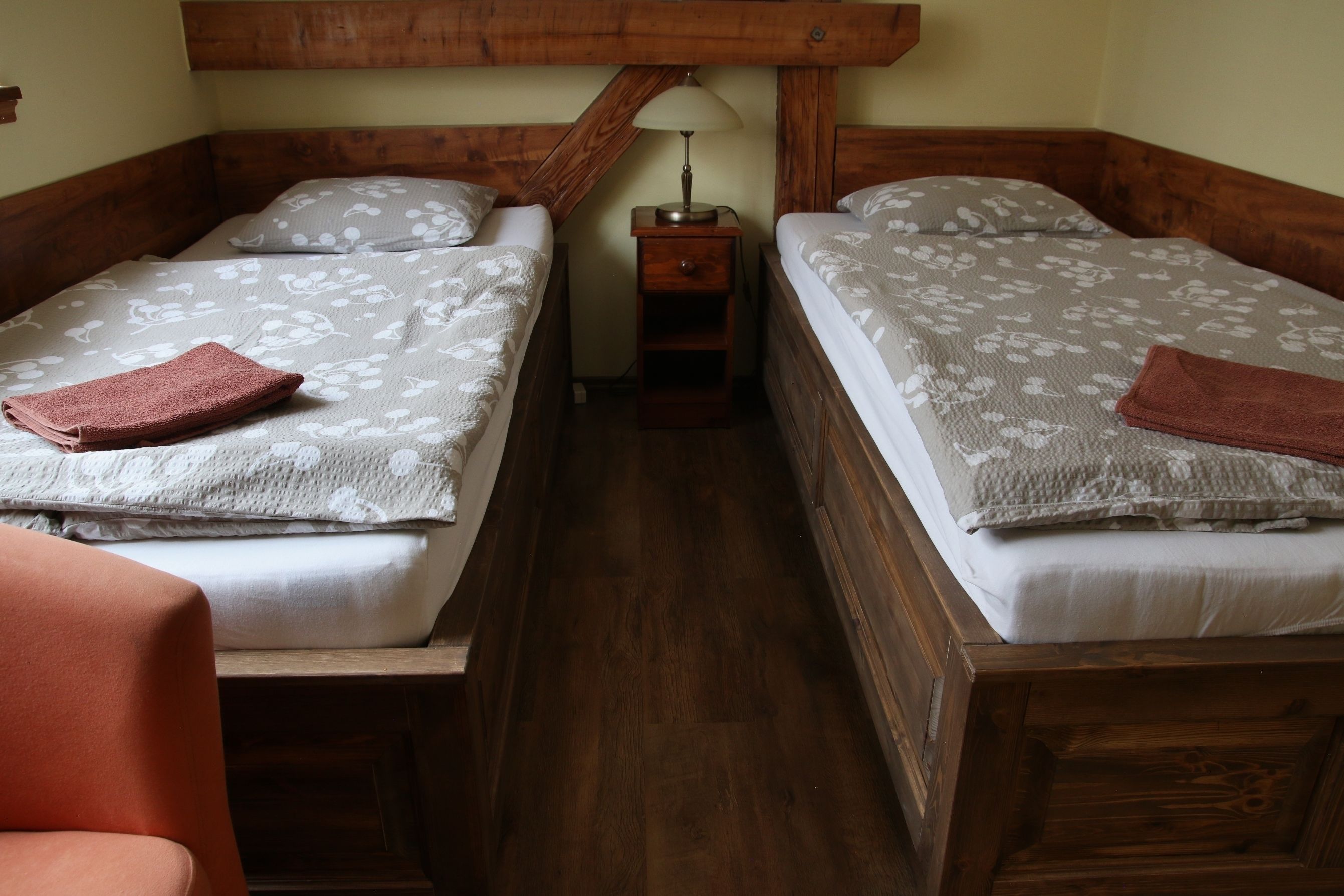 Una habitación con dos camas de madera, cada una con una almohada y una toalla doblada. Entre las camas hay una mesita de noche de madera con una lámpara.
