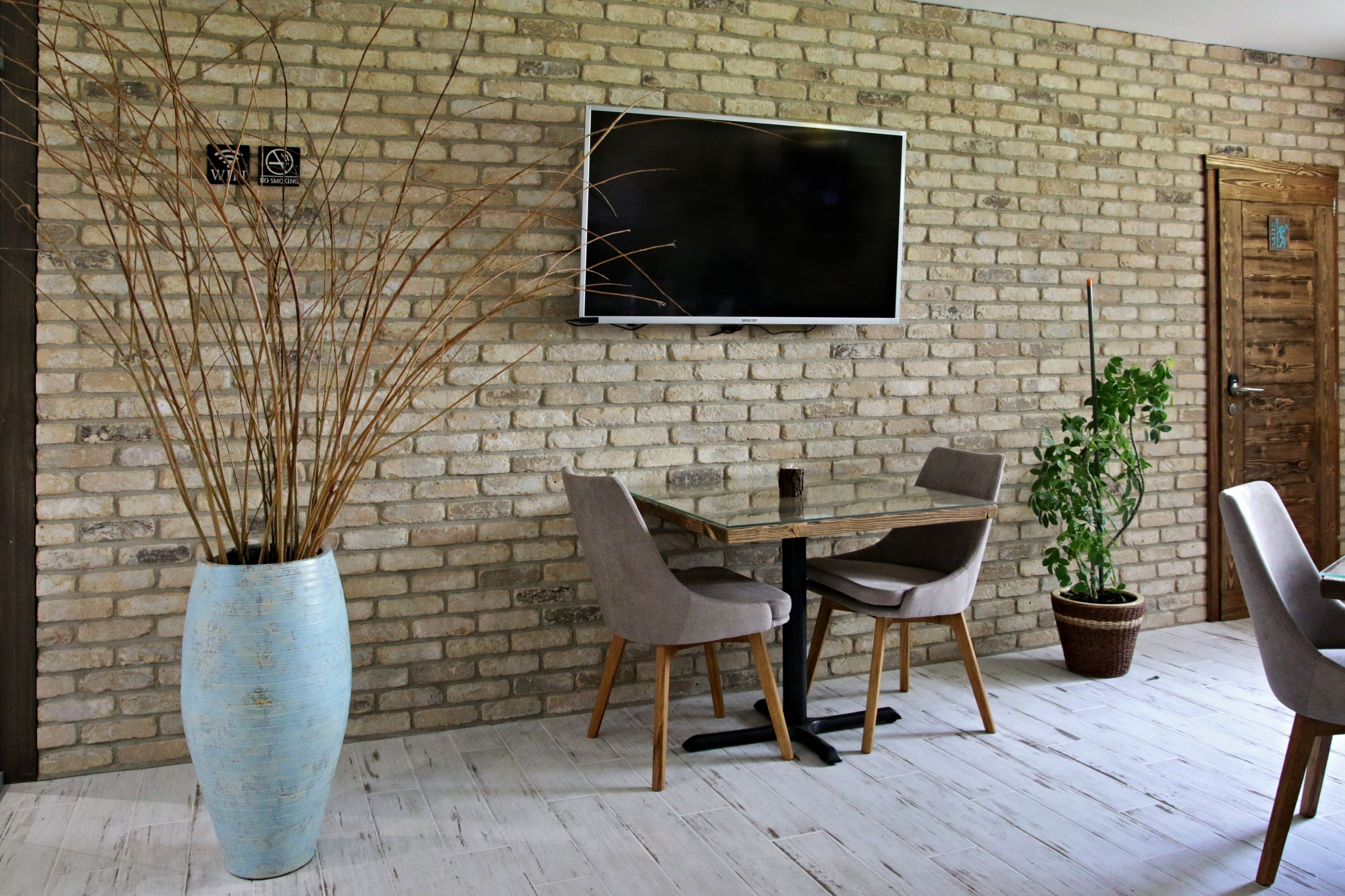 A dining area with a brick wall, a flat-screen TV, a large vase with branches, and a potted plant.
