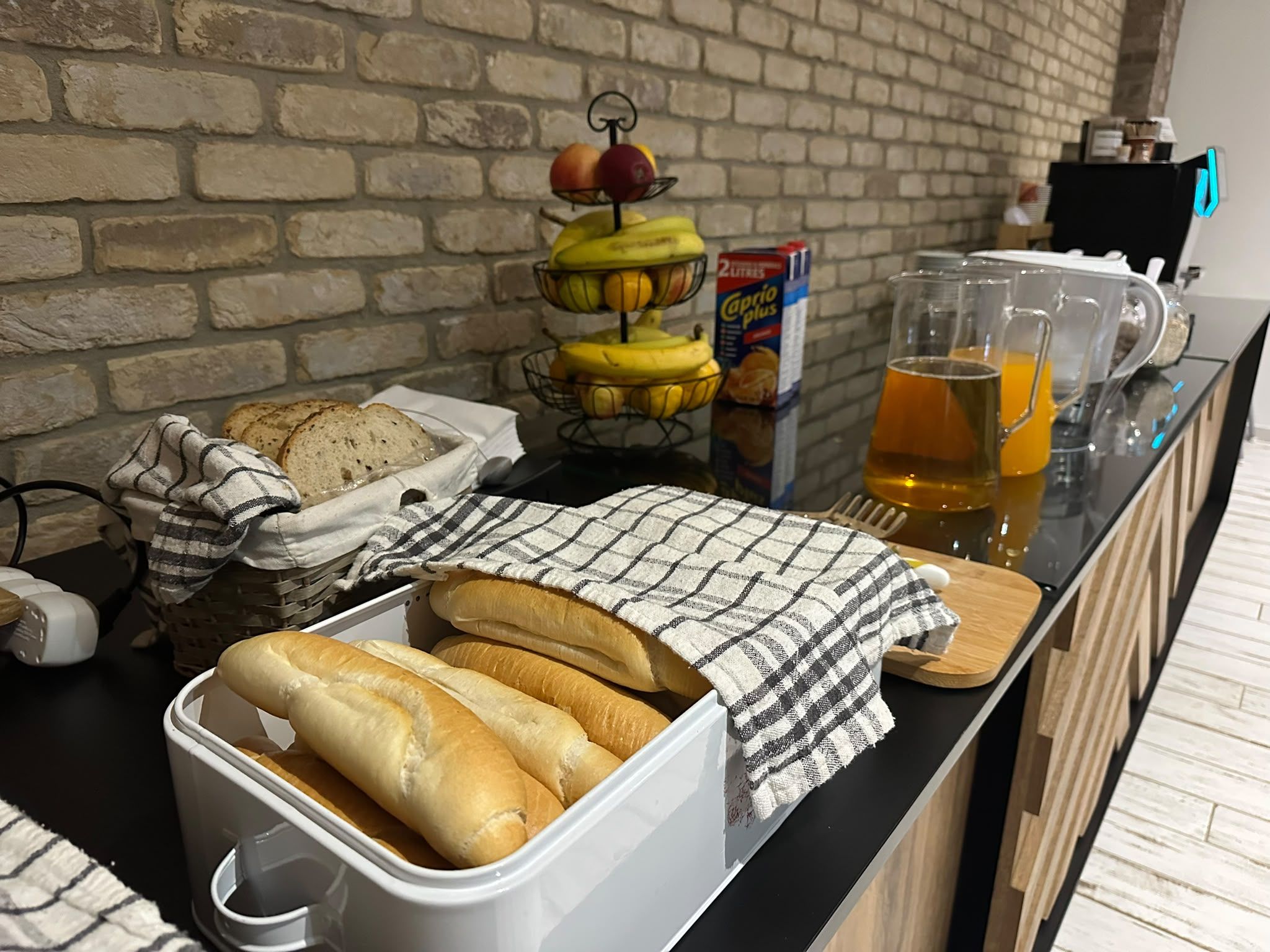 Desayuno con pan, una cesta de frutas, una caja de cereales y una jarra de jugo de naranja sobre una encimera de cocina.