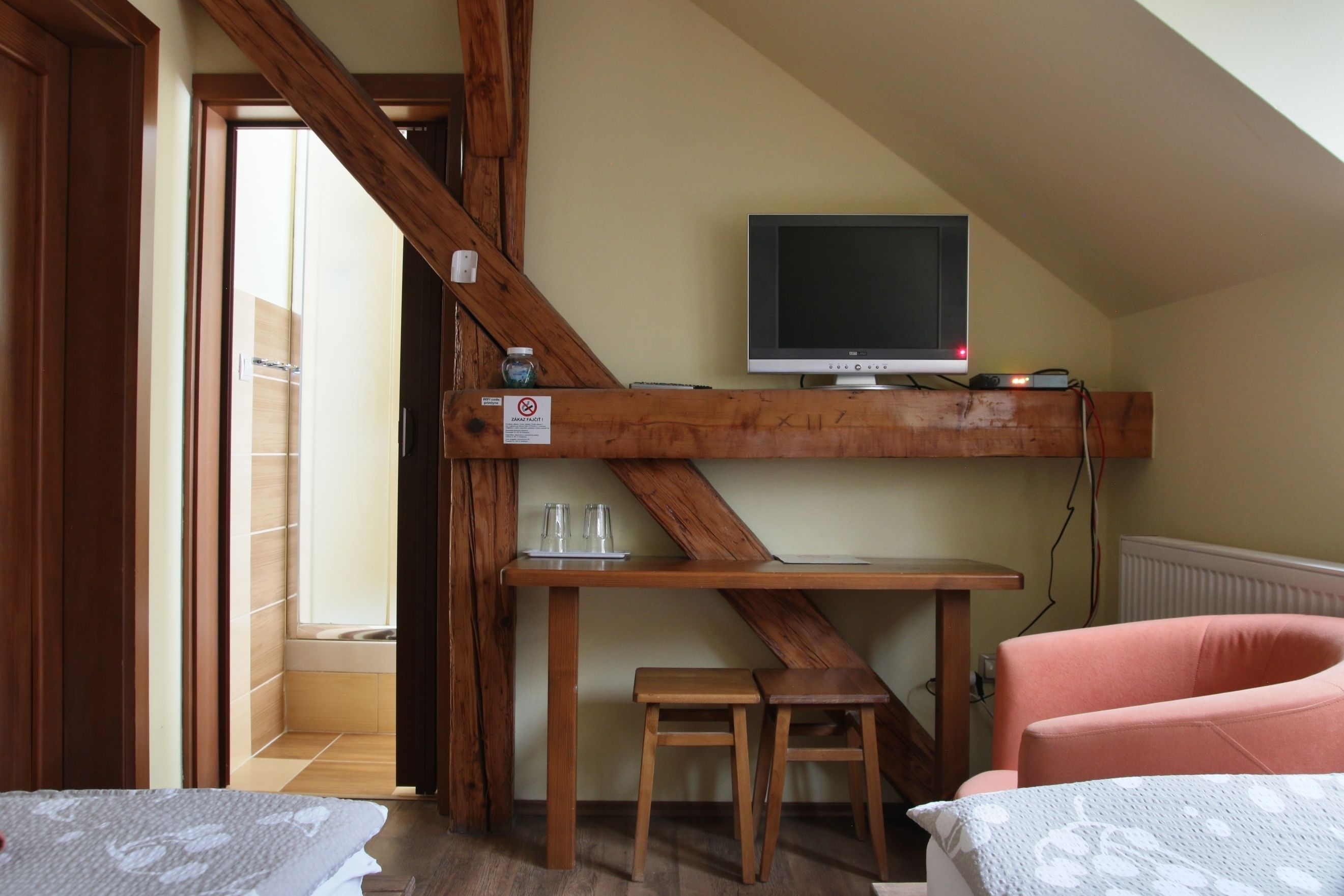 A room with a wooden ceiling, a television on a shelf, a small table with glasses, a pink chair, and a bed with a patterned bedspread.