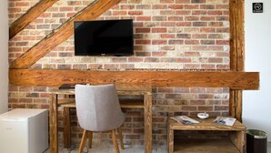 Una habitación con pared de ladrillo, un televisor de pantalla plana montado, un escritorio de madera con una silla, una mesita con revistas y un mini refrigerador blanco.