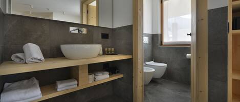A modern bathroom with a large white basin, wooden shelves, a toilet, and a window.
