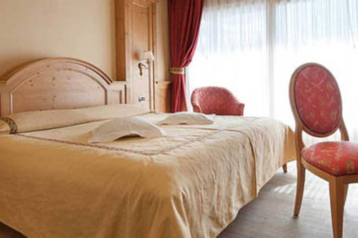 A hotel room with a large bed, a wooden headboard, a nightstand with a lamp, a red chair, and a window with curtains.