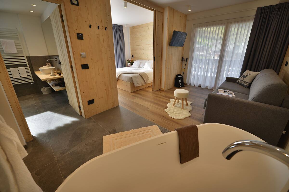 A modern hotel room with a wooden floor, a large bed, a sofa, a TV, and a bathroom with a bathtub.