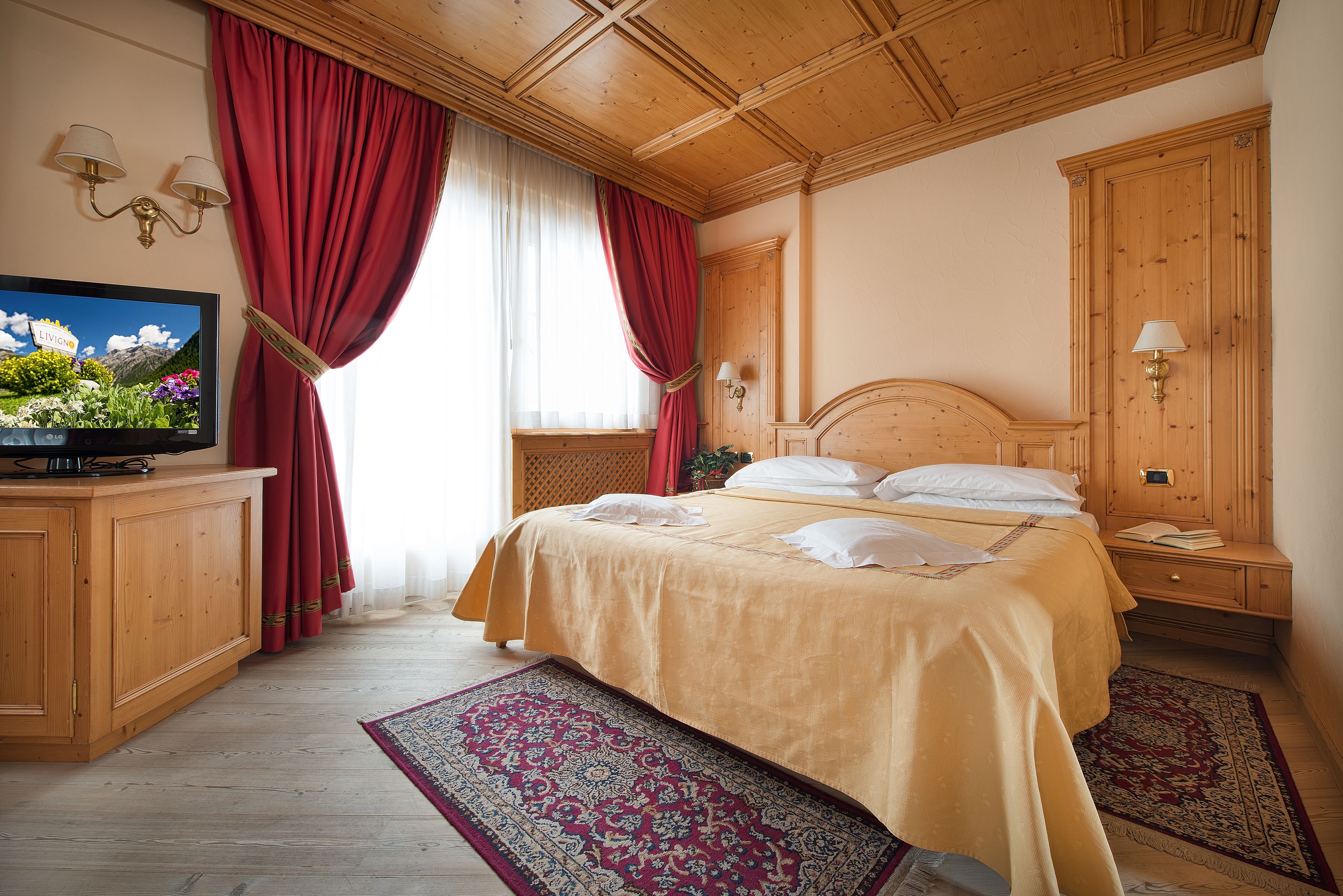 Una habitación de hotel con cama, armario de madera, televisión y ventana con cortinas rojas.