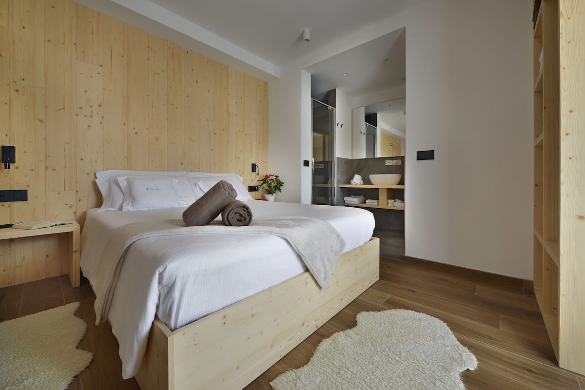 A bedroom with a wooden bed, a bedside table, a white rug, and a bathroom visible through an open door.