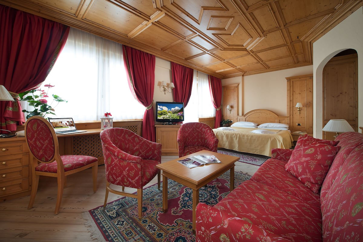 A room with a wooden ceiling, red curtains, a bed, a desk with a computer, a TV, and a red patterned sofa.