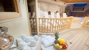 A dining area with a round table set for a meal, a fruit basket, and a view of a pool area with a wooden deck and a railing. - Hotel Valtellina (Livigno)