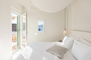 A bedroom with a bed, pillows, a nightstand, a window with a view, and a door leading to an outdoor area. - Eneos Kythnos Beach Villas (Kithnos)