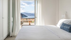 A bedroom with a bed, a nightstand, a lamp, and a view of the sea and mountains. - Eneos Kythnos Beach Villas (Kithnos)