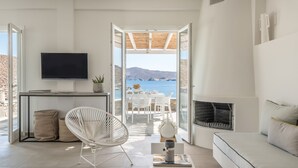 A modern living room with a TV, white furniture, a fireplace, and a view of the sea. - Eneos Kythnos Beach Villas (Kithnos)